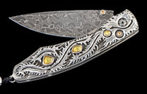 Ornate folding knife with a patterned Damascus blade and a sterling silver handle featuring intricate scrollwork and yellow gemstones.