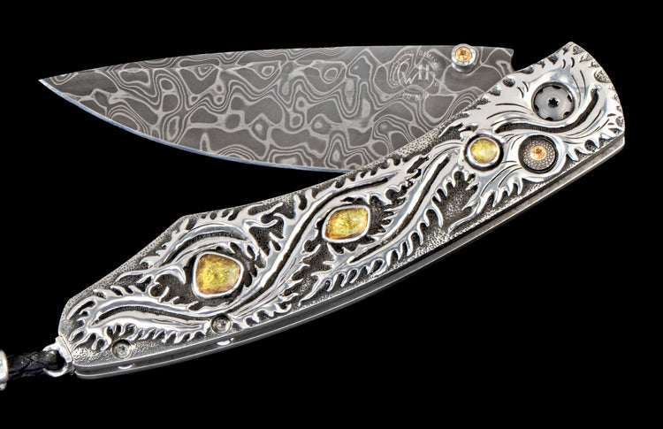 Ornate folding knife with a patterned Damascus blade and a sterling silver handle featuring intricate scrollwork and yellow gemstones.