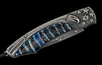 Ornate folding knife with an engraved silver handle, featuring an eagle, stars, and blue-green striped inlay, with a Damascus steel blade.
