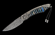 Folding knife with a Damascus steel blade, silver eagle and star handle with blue gemstones, colorful striped scales, and a black braided lanyard.