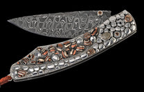 Ornate folding knife with a Damascus steel blade, textured handle inlaid with silver-toned pebbles, striped stones, and a braided lanyard.