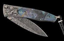 Partially open folding knife with a silver Damascus blade and iridescent blue-green abalone handle, featuring an eagle, stars, and small brown gems.