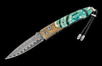 Ornate folding knife with a wavy Damascus blade, gold-engraved metal bolsters, and swirling teal handle scales, featuring a black braided lanyard.
