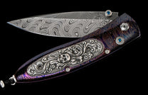 Open Damascus steel knife with purple and blue swirled handle, ornate silver skull design, and blue gemstones.