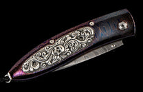 Custom folding knife with a dark iridescent purple and blue patterned handle, featuring an ornate silver skull inlay, and a Damascus steel blade.