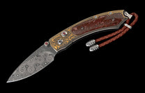 Custom folding knife with a swirling Damascus blade, intricate gold engraving, red gemstone accents, reddish-brown handle scales, and a braided leather lanyard.