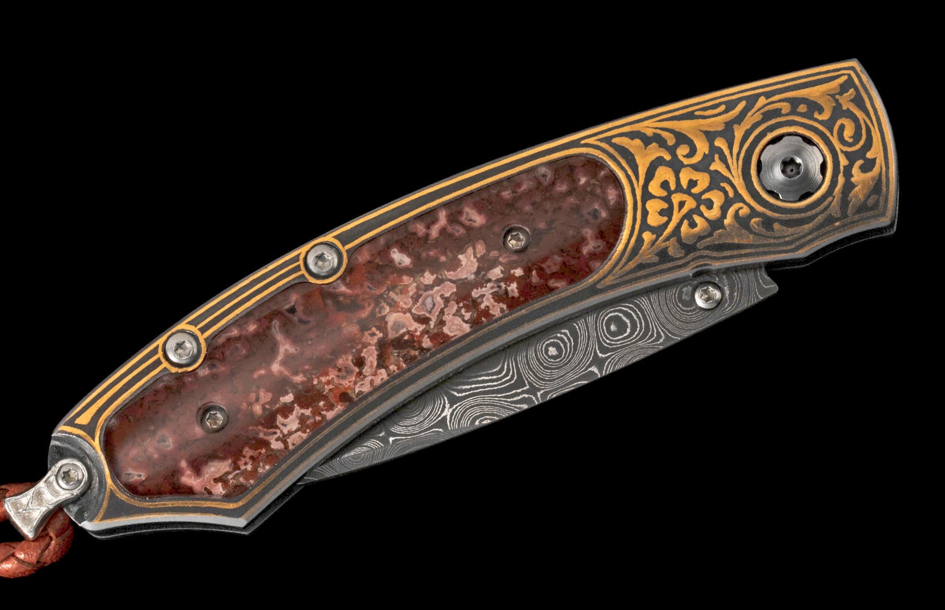 Decorative folding knife with dark handle, gold scrolling, red stone inlay, and Damascus steel blade, with a braided leather lanyard.