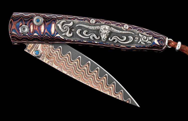 Multicolored wavy patterned folding knife with blue gems, a silver bull skull inlay, and a wavy Damascus blade, partially open.