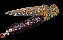 Partially open folding knife with wavy Damascus blade, multi-colored layered handle, gold-engraved accents with blue gemstones, and braided leather lanyard.