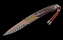 Ornate folding knife with wavy Damascus blade, gold floral engraved bolsters, two blue gemstones, and multicolored patterned handle scales, with a braided leather lanyard.