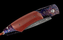 Closed folding knife with a speckled red handle, blue and purple lightning-patterned bolsters, and a Damascus steel blade, with a brown leather lanyard.