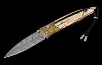 Folding knife with a swirling Damascus blade, ornate gold-engraved bolster, patterned ivory-like handle scales, and a black braided lanyard.