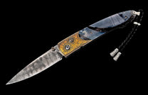 Ornate folding knife featuring a swirling Damascus blade, gold floral engraved handle, dark blue inlay, and black lanyard with silver beads.