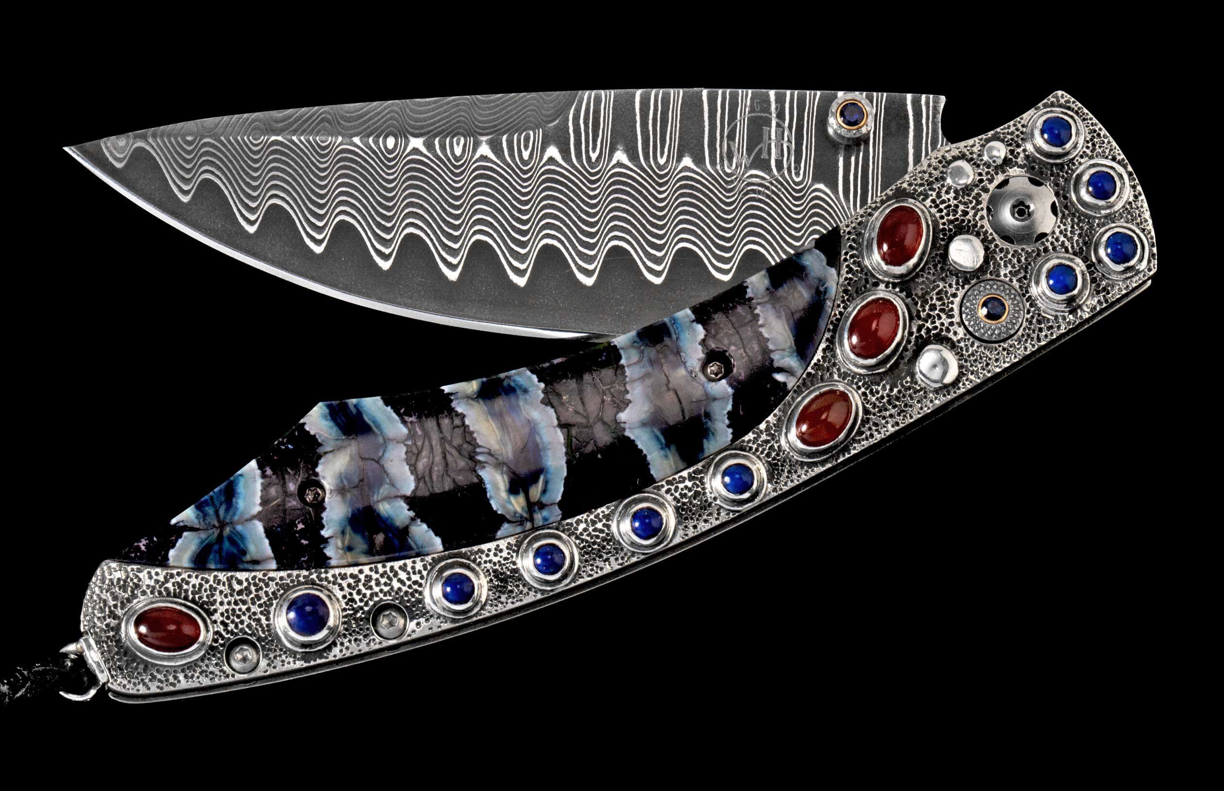 Ornate folding knife with wavy Damascus steel blade and a textured handle adorned with red and blue stones and a striped inlay.