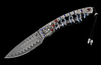 Ornate knife with a wavy Damascus blade, textured silver handle featuring red and blue gemstones, marbled shell grip, and braided cord.