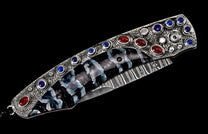 Ornate folding knife with textured silver-toned handle, blue and red gemstones, patterned inlay, and Damascus blade.