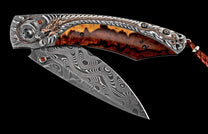 Folding knife, partially open, with a wavy Damascus steel blade and ornate silver handle featuring a bronze dragon, burled wood, red gems, and a leather lanyard.