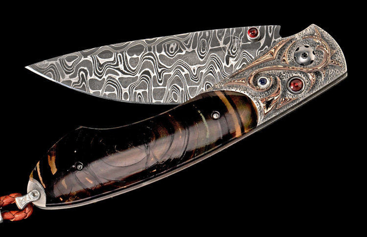 Damascus steel folding knife with a wavy blade, iridescent brown handle, ornate metal frame with swirling inlays and colorful gemstones.
