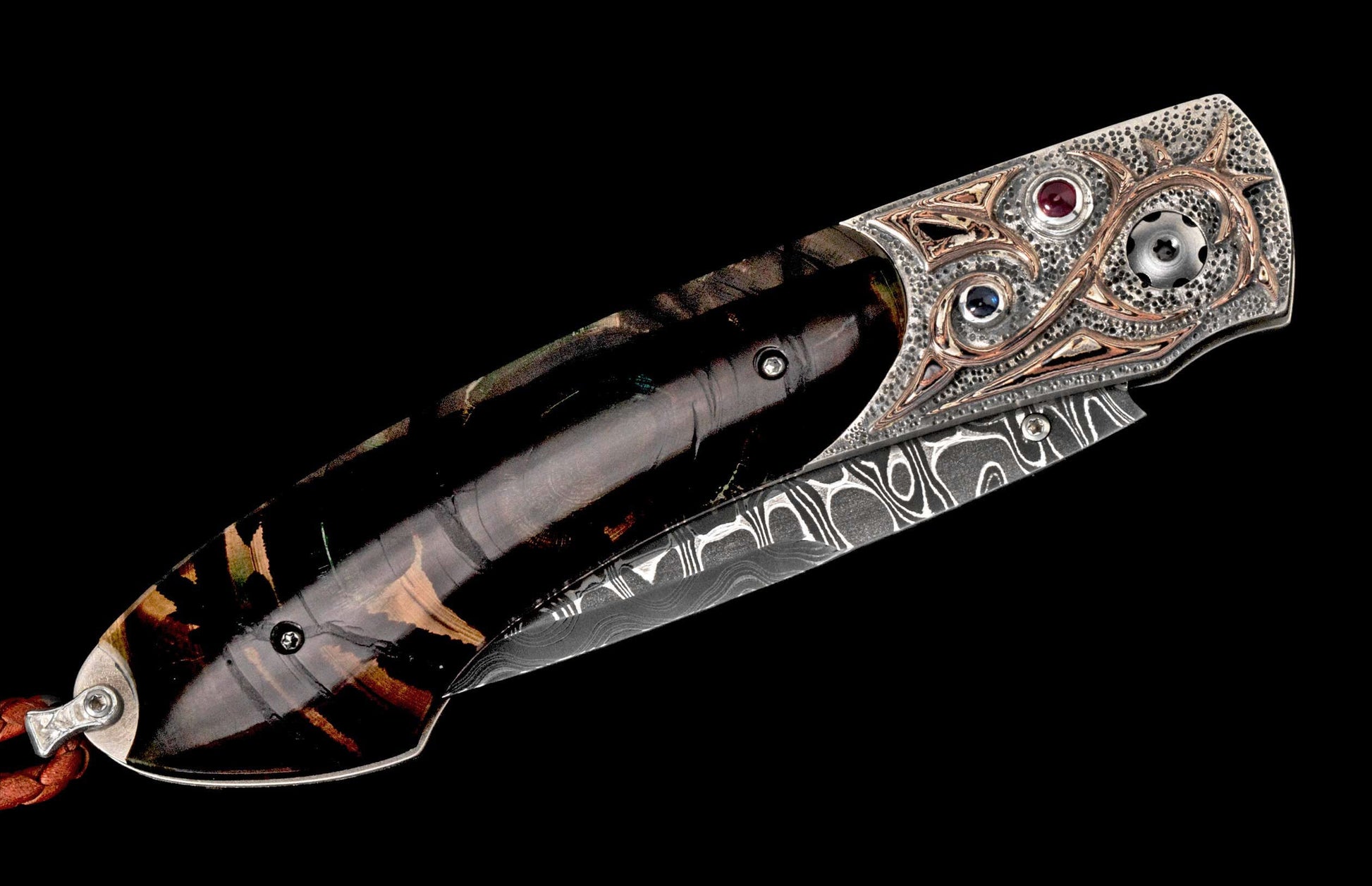 Artisan folding knife with a dark patterned handle, ornate silver and bronze bolster with red and blue gems, and a Damascus steel blade.