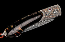 Artisan folding knife with a dark patterned handle, ornate silver and bronze bolster with red and blue gems, and a Damascus steel blade.