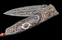 Decorative folding knife with a patterned Damascus steel blade, a layered black, white, and red handle, intricate bronze detailing, and red gem inlays.