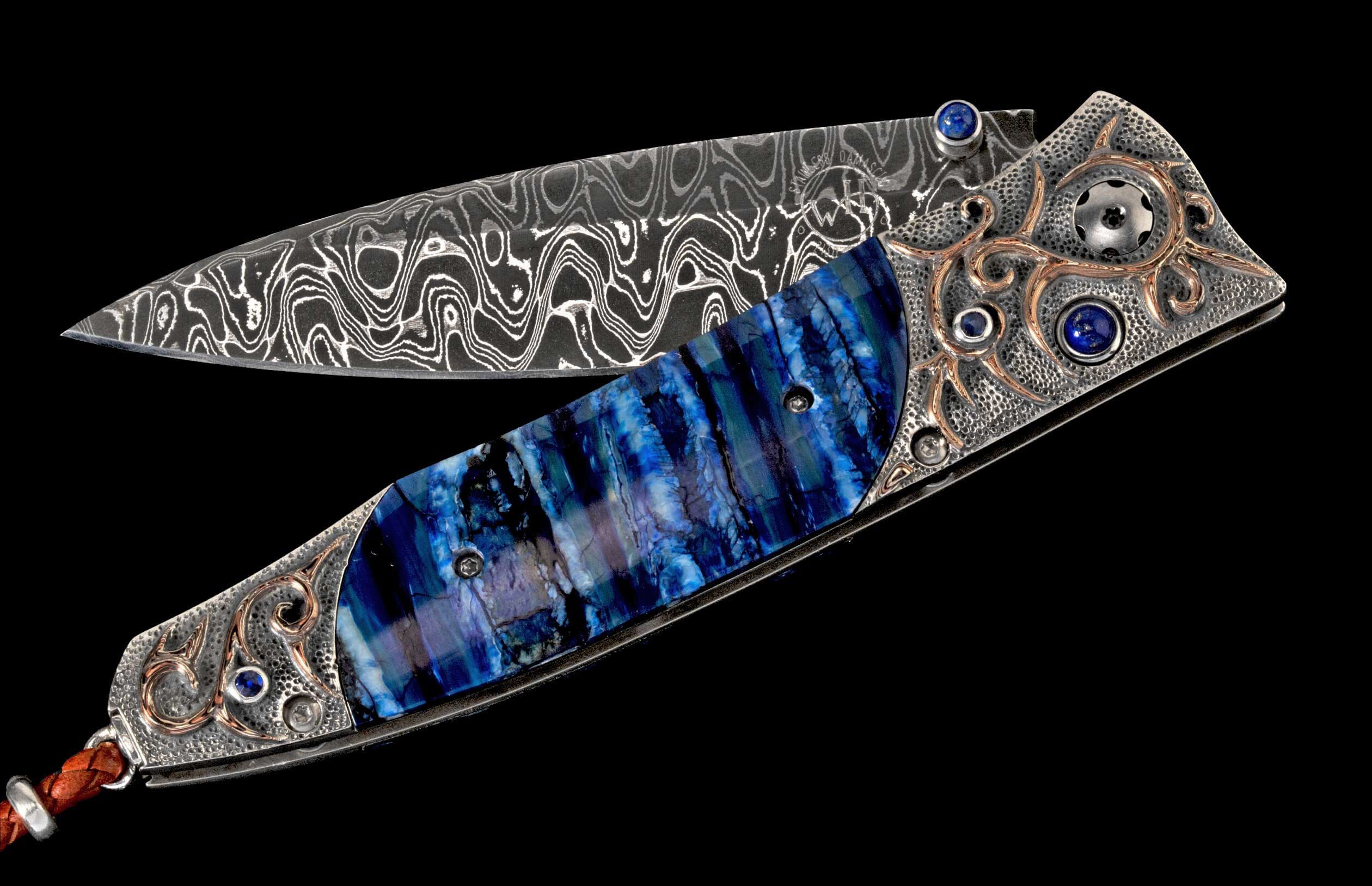 Ornate folding knife with wavy Damascus steel blade, vibrant blue stone handle, silver frame, gold scrollwork, blue gems, and leather lanyard.