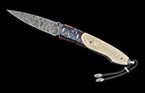 Folding knife with a patterned Damascus blade, a vibrant swirled handle featuring blue gems, and a carved off-white scorpion handle with a black leather lanyard.