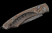 Ornate folding knife featuring a textured silver frame with dark scales, intricate bronze inlays, a Damascus blade, and blue and red gemstones.