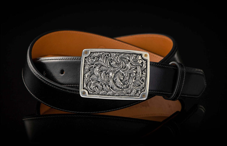Silver-toned engraved buckle on a black leather belt with an orange underside, coiled on a reflective surface.