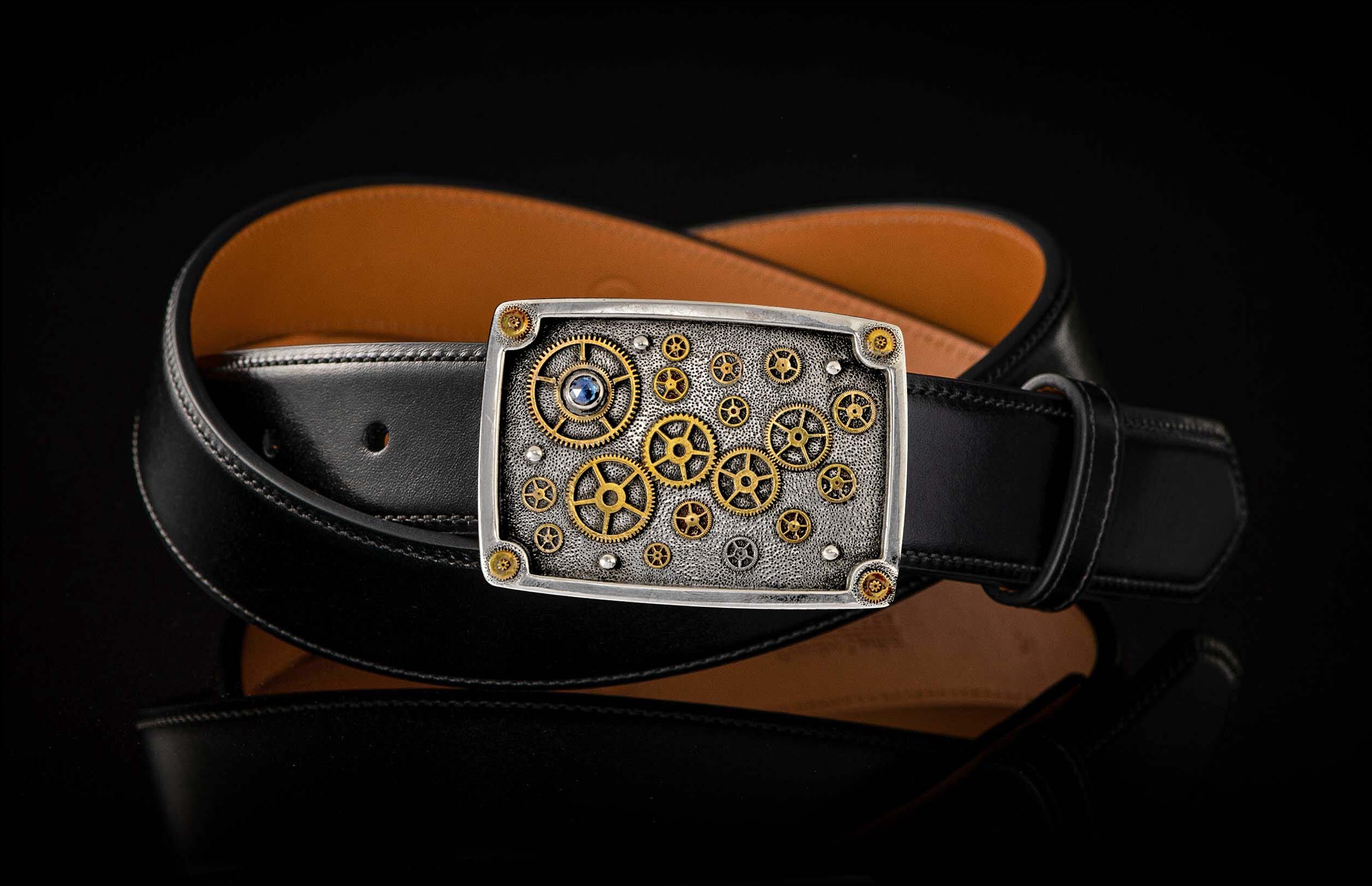 Rectangular buckle with silver-toned textured metal, brass gears, and a blue gem, attached to a black leather belt.