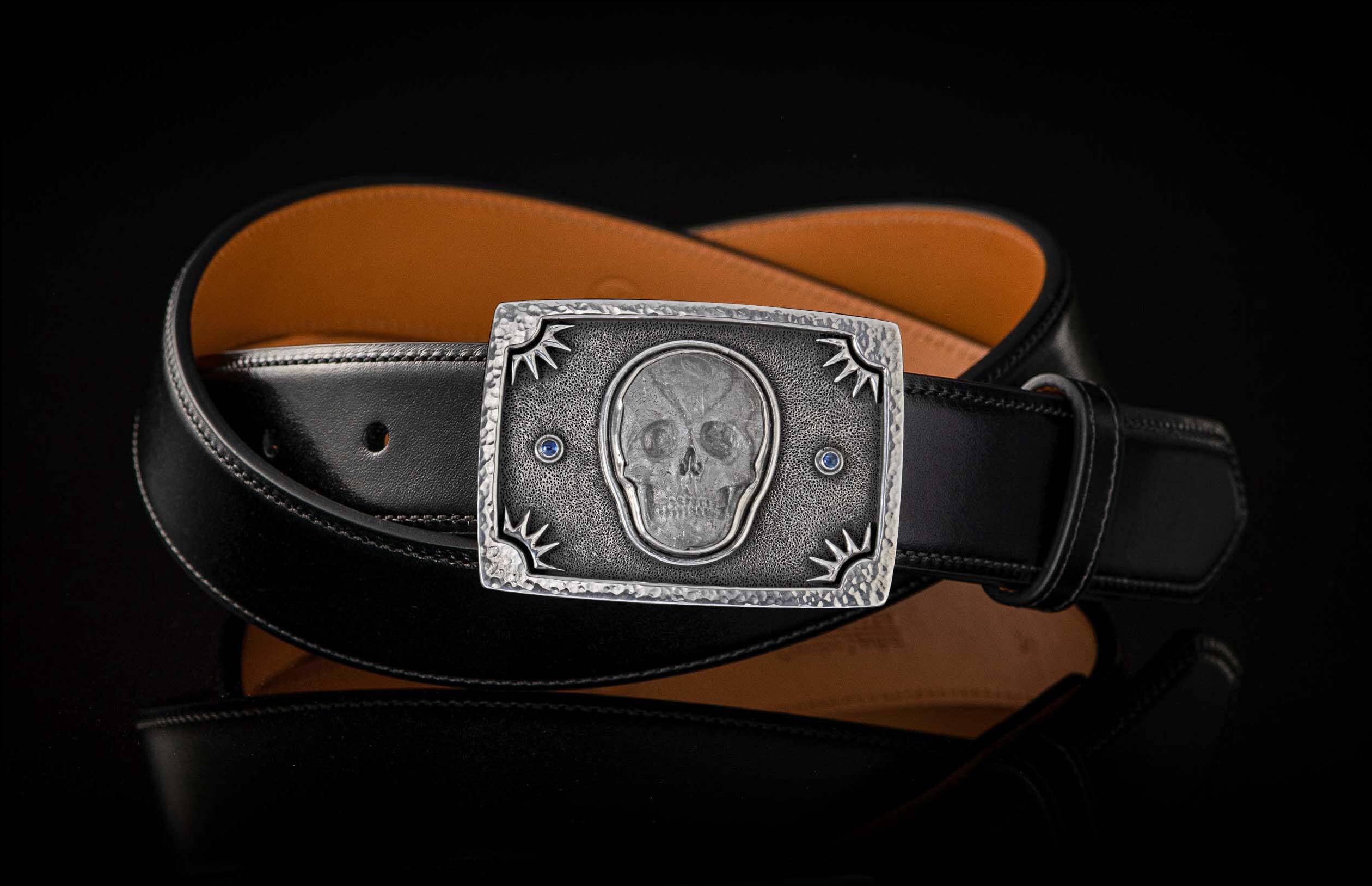 Silver hammered buckle with a skull design, blue accents, and starburst details, on a black leather belt.