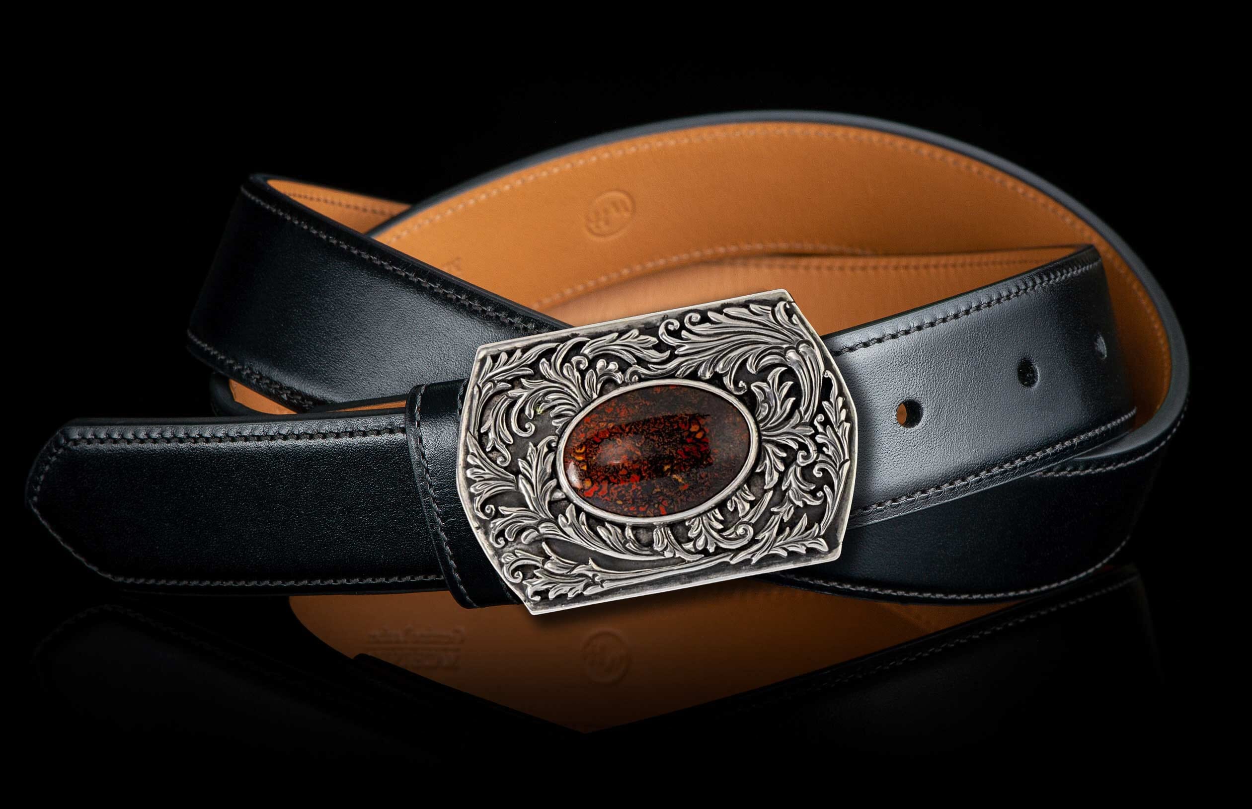 Silver-toned buckle with intricate scrollwork, featuring an oval reddish-brown patterned stone, attached to a black leather belt with tan lining.