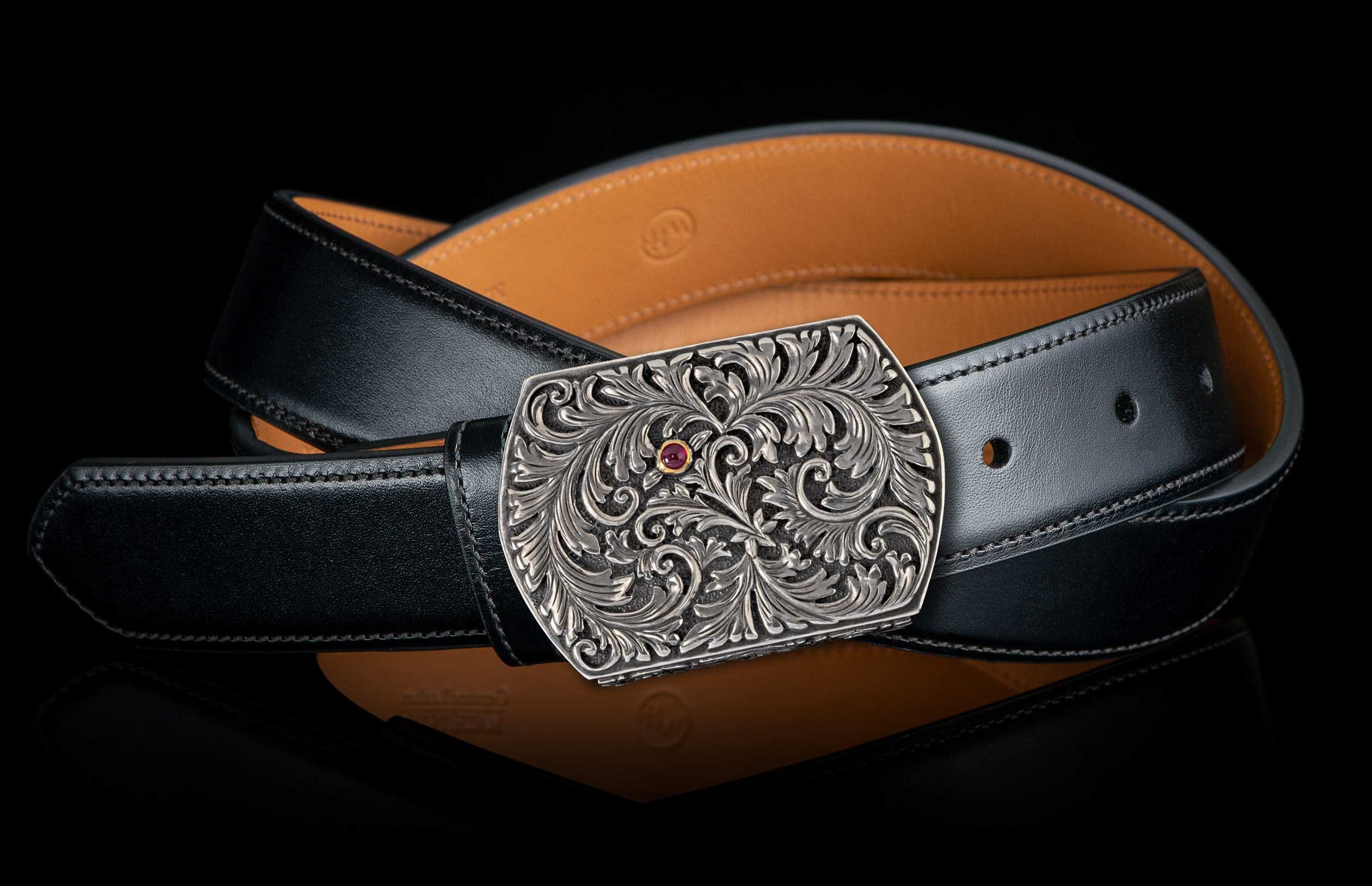 Silver-toned buckle with intricate scrollwork and a red gem, attached to a black leather belt.
