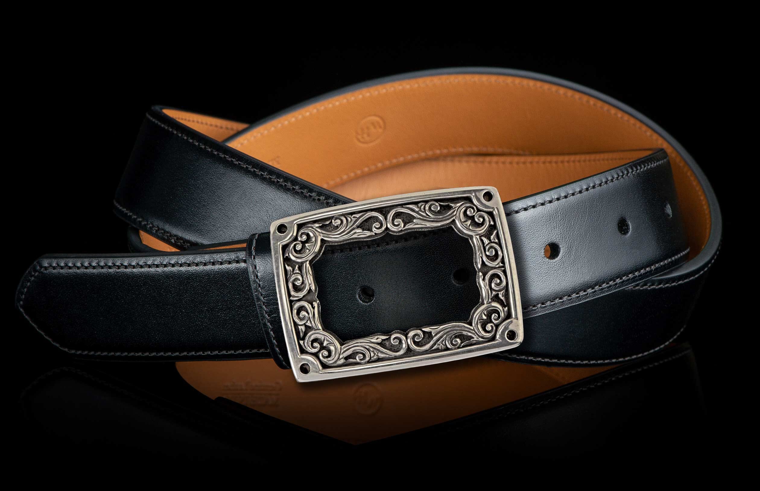 Ornate silver-tone rectangular buckle with filigree detail on a black leather belt with tan lining.