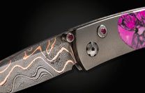 Damascus steel folding knife with rose gold wave pattern and pink gemstone accents, side view close-up