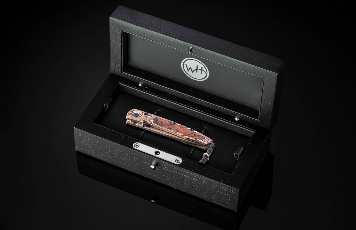 Luxury folding knife with rich riverwood handle by William Henry, displayed in black presentation box with silver hardware and braided lanyard.