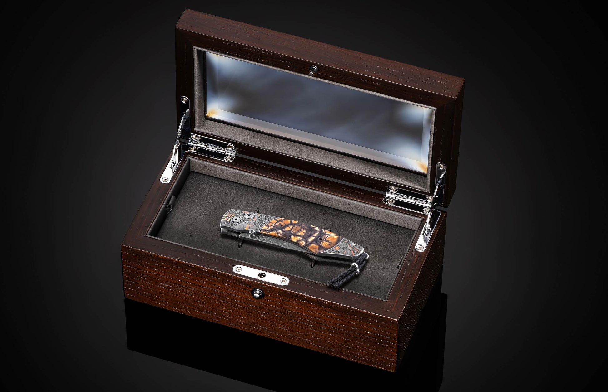 Luxury folding knife with amber and black mosaic handle, by B12, displayed in dark wood presentation box with glass lid, top-down view