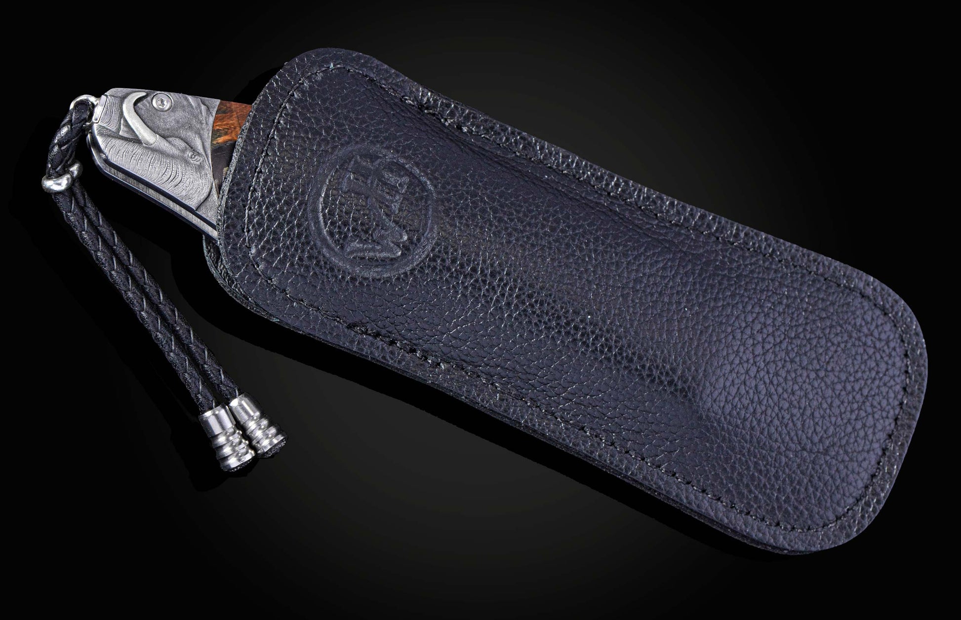 Folding knife with an embossed boar head handle, wood inlay, and braided lanyard, partly in a black textured leather sheath with an embossed logo.