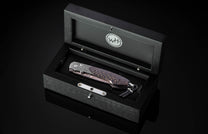 Black folding knife with dark wood grain handle and Damascus steel blade, by B12, displayed in luxury black presentation box with silver accents