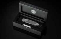 Grey folding knife with a textured carbon fiber handle and Damascus blade, displayed inside an open black wooden presentation box with a silver WH logo in the lid.