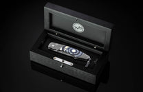 Luxury folding knife with blue and silver marbled handle by B12, displayed in black wooden box with engraved logo, top-down view