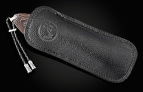 Knife with wavy patterned handle and silver-toned bolster, partially sheathed in black textured leather with an embossed circular logo, featuring a braided leather lanyard.