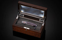 Folding knife with a dark patterned handle, orange accents, and a blue lanyard, displayed in an open dark brown wooden case with a clear lid.