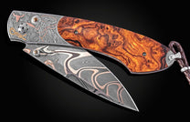 Damascus steel folding knife with burl wood handle and engraved cowboy motif by B12, featuring rose gold accents and braided leather lanyard, side view