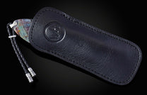 Knife with colorful veined handle partially in a black leather sheath with embossed logo and braided lanyard.