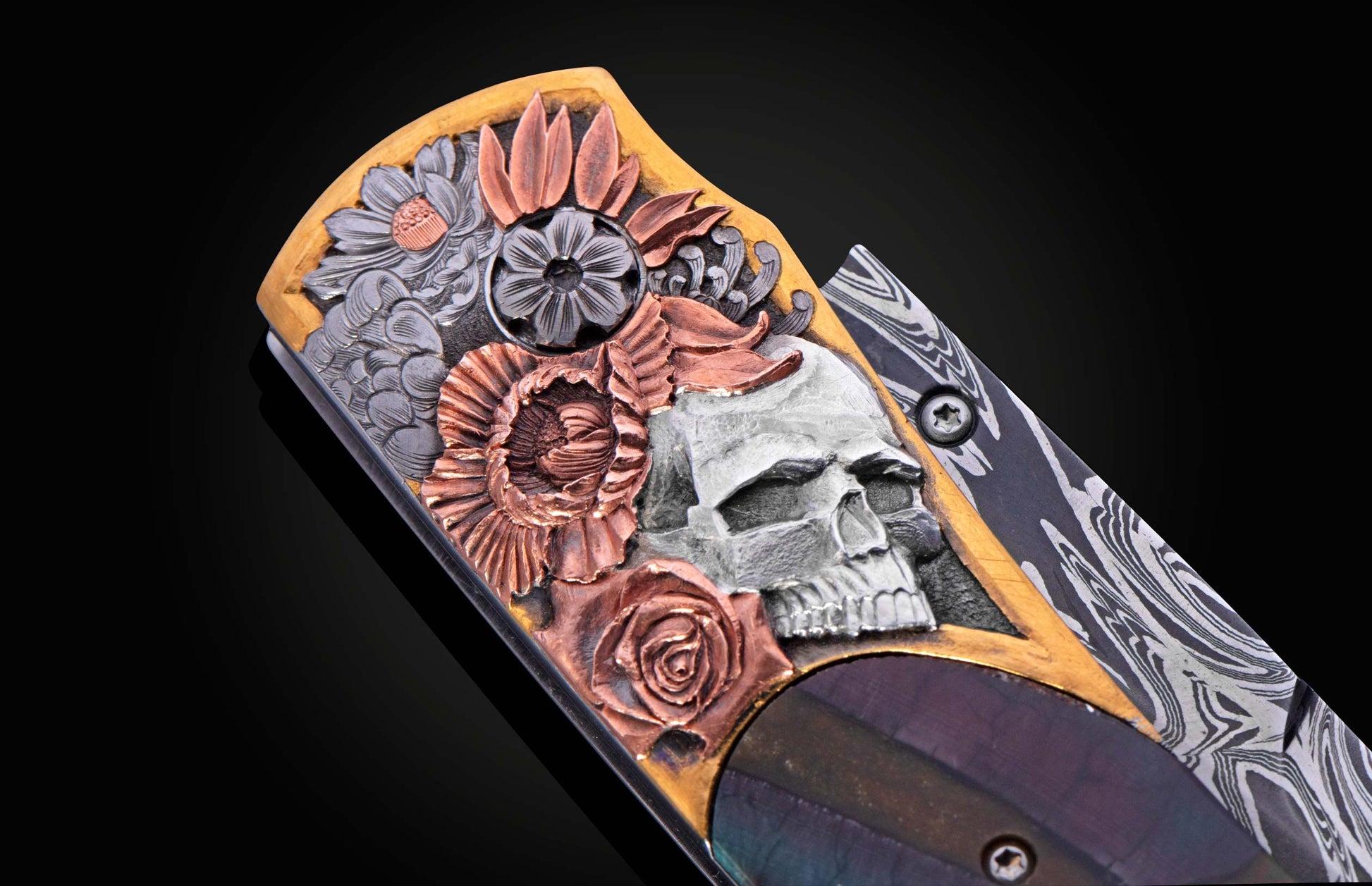 Ornate knife with a handle engraved with a silver skull, copper flowers, gold border, and dark iridescent inlay, featuring a damascus steel blade.