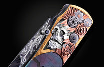 An ornate knife with a detailed silver skull, copper and silver floral carvings, gold accents, a Damascus steel blade, and black gemstones.