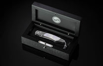 Gilded Ghost folding knife with damascus steel handle and mother-of-pearl inlay by B12, displayed in black wooden presentation box with silver hardware, top-down view