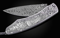 Damascus steel folding knife by B12 with intricate Halloween-themed engraving, silver-toned handle, and black gem accents, displayed open on black background