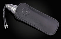 Silver engraved folding knife with Halloween-themed skeleton design, by B12, in black leather sheath with braided cord and metal tips, side view.